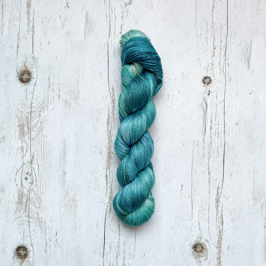 A single hank of variegated yarn in shades of teal, blue, and green, laid flat on a distressed white wooden surface.