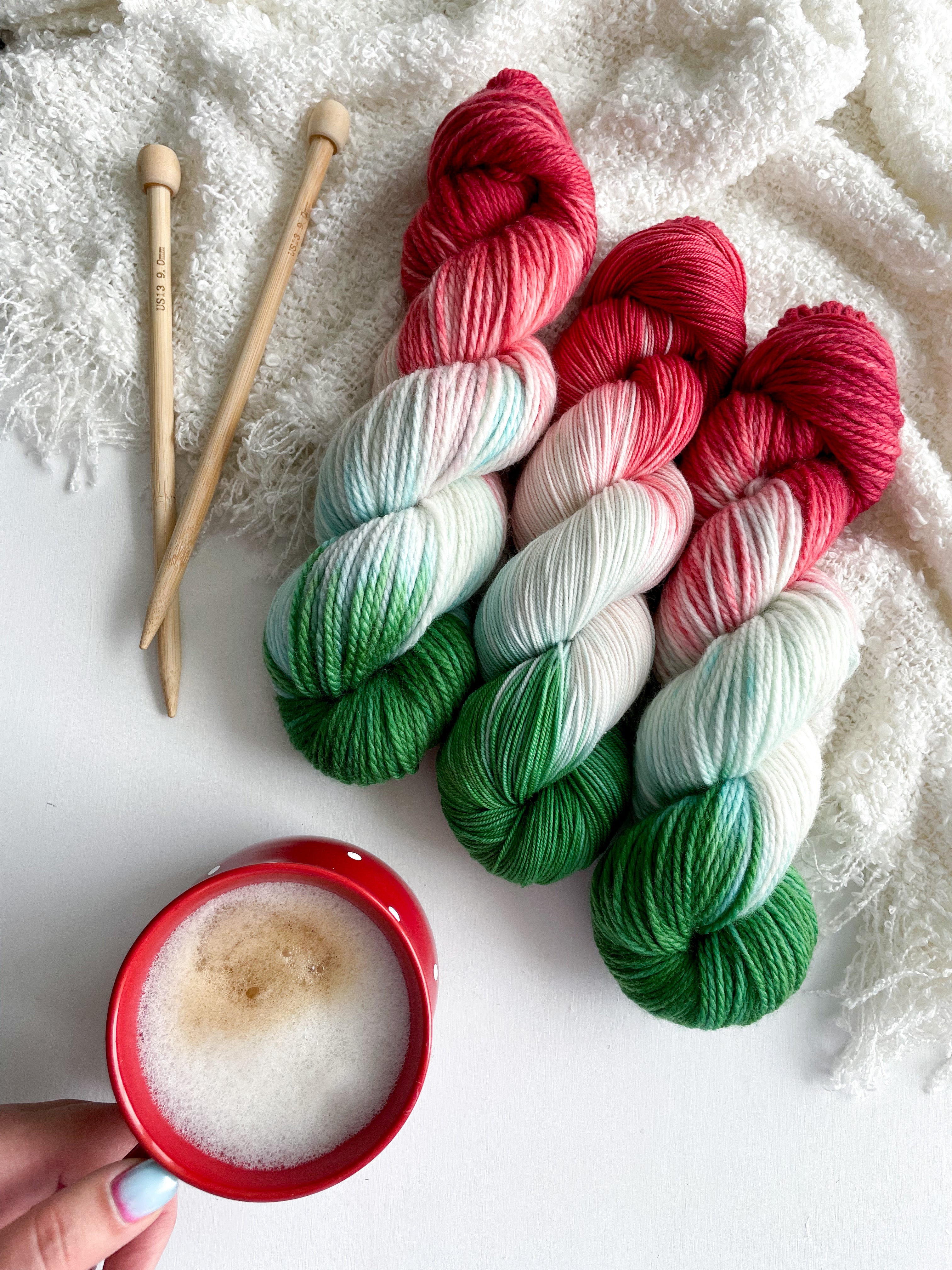 Three skeins of red, white, and green hand-dyed yarn with knitting needles and a coffee cup.