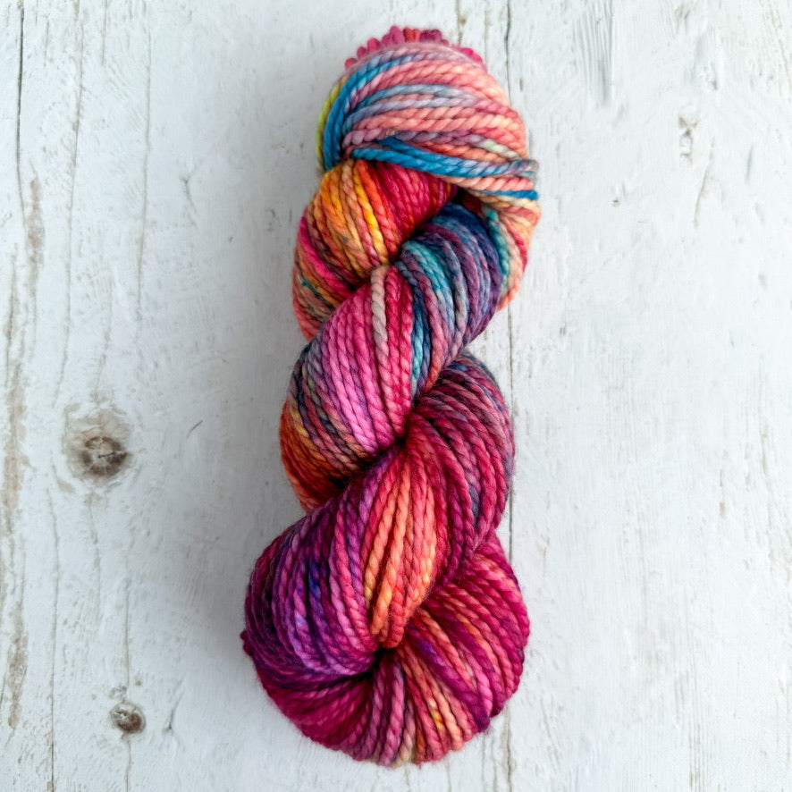 A single skein of variegated yarn with vibrant pink, orange, yellow, blue, and purple tones, displayed on a white-washed wooden surface.