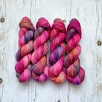 Four hanks of variegated yarn laid flat on a whitewashed wooden surface. The yarn displays a rich blend of magenta pink, purple, orange, and hints of blue, showcasing its variegated color story.