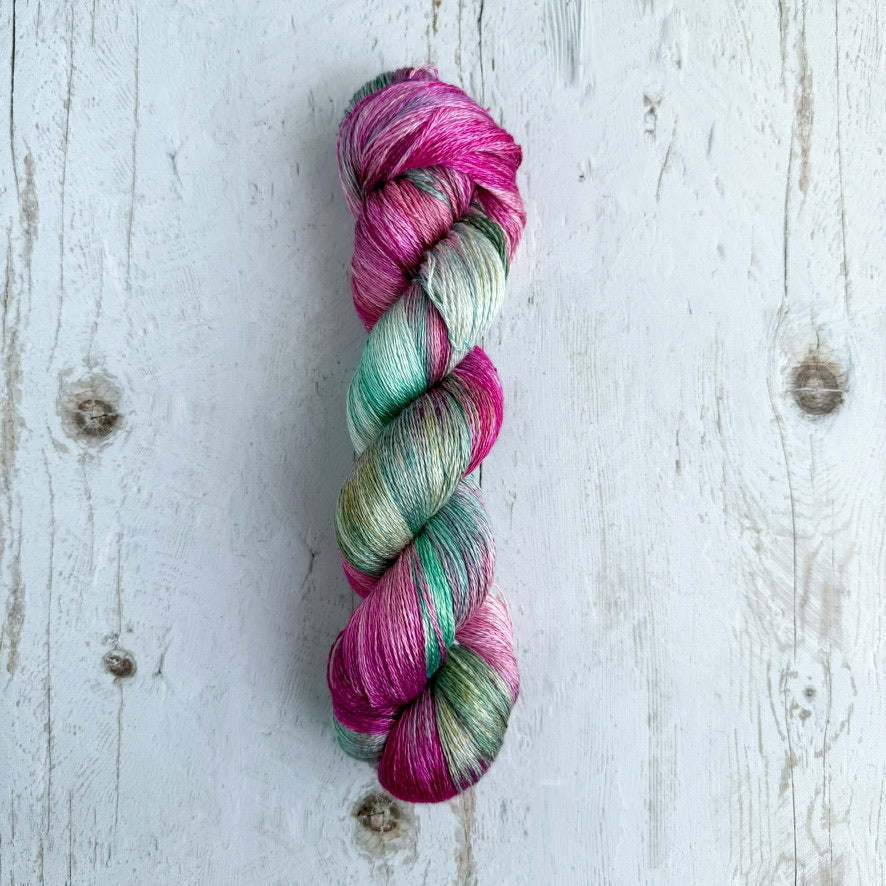 A single skein of variegated yarn with pink, mint green, and gray-blue tones, displayed vertically on a whitewashed wooden surface.